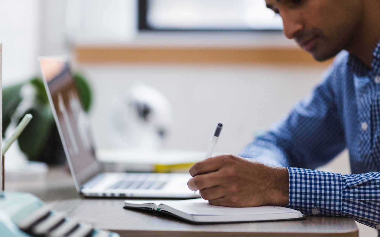 Person taking notes beside a laptop in a focused office workspace.